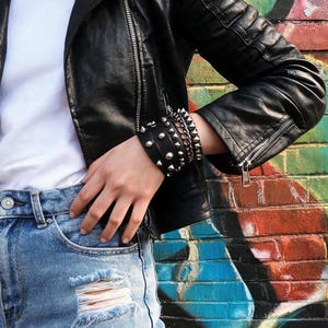 Op de afbeelding: Een zwart leren motorjack met zilveren ritsen en doorgestikte schouderdetails, gedragen over een effen wit t-shirt en lichtblauwe distressed denim shorts, voorzien van een brede zwarte leren polsband met zilveren metalen studs.