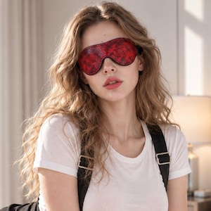 Könnte beinhalten: Eine rote Schlafmaske mit Schlangenledermuster und einer glatten, glänzenden Oberfläche. Die Maske hat eine konturierte Form, die die Augen bedeckt, und ein dünnes schwarzes Gummiband an den Seiten, um sie sicher am Kopf zu befestigen.