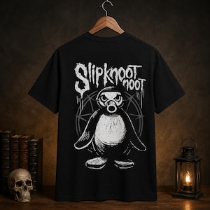 May include: A black short-sleeve t-shirt featuring a white graphic print of a penguin wearing a gas mask. Above the penguin, the text reads Slipknot Noot in a distressed, stylized font. A circular geometric design sits behind the penguin.