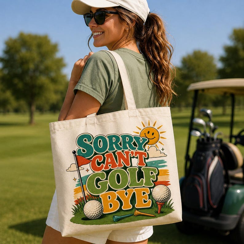 A cream-colored canvas tote bag featuring a colorful graphic print with the text Sorry Can't Golf Bye. The design includes a yellow sun, a golf ball on a tee, a red flag, and green grass elements set against a retro-style background.
