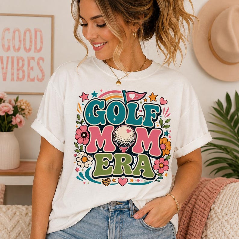 A white short-sleeve t-shirt featuring a colorful graphic print that reads GOLF MOM ERA in large, bold, multi-colored letters. The design includes a golf ball, a flag, flowers, stars, and hearts in teal, pink, and green.