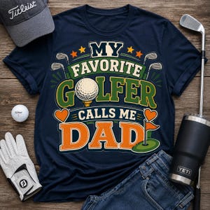 A navy blue t-shirt featuring a graphic with the text "MY FAVORITE GOLFER CALLS ME DAD". The design includes a golf ball on a tee, golf clubs, hearts, and a golf flag. The shirt is surrounded by a golf cap, a golf ball, a glove, and a club.