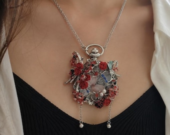 May include: A silver-toned pocket watch pendant necklace featuring an ornate floral arrangement with red roses, small white pearls, and delicate butterfly accents. The watch face is partially obscured by the decorative embellishments and two hanging chains.