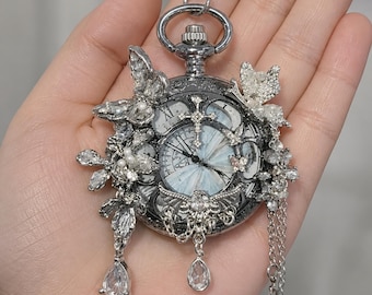 May include: A silver-toned pocket watch pendant featuring a light blue clock face with Roman numerals. The watch is adorned with intricate silver butterfly motifs, clear crystal accents, and three dangling teardrop-shaped clear crystal charms.