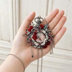 Puede incluir: Un colgante de reloj de bolsillo de metal oscuro con esfera blanca, números romanos e ilustración de mariposa. El reloj está adornado con rosas rojas, pequeñas perlas blancas, cristales rojos y cadenas plateadas con un dije de gota negro.