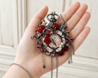 May include: A dark metal pocket watch pendant featuring a white clock face with a butterfly illustration, adorned with red roses, small red crystals, two white pearls, and dangling silver chains with a black teardrop charm and clear crystals.