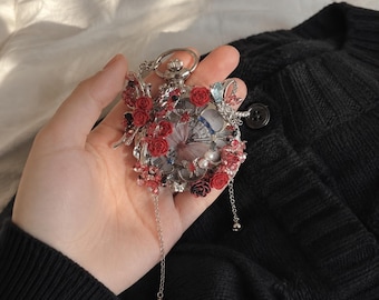 May include: A silver-toned pocket watch adorned with red floral accents, clear crystals, and small white pearls. The watch face features a butterfly graphic, and two delicate silver chains with small spherical charms hang from the bottom edge.