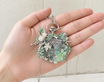 May include: A silver-toned pocket watch pendant featuring a circular face with black hands and numerals. The exterior is adorned with green and white floral embellishments, small clear crystals, and two delicate silver chains with teardrop charms.