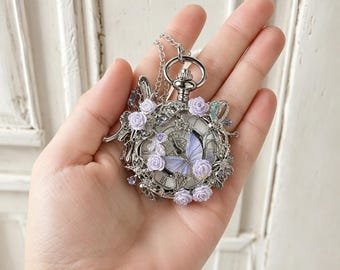 May include: A silver-toned pocket watch pendant featuring a white clock face with black Roman numerals. The exterior is decorated with light purple miniature roses, a central light purple butterfly, and small clear crystals on a silver chain.