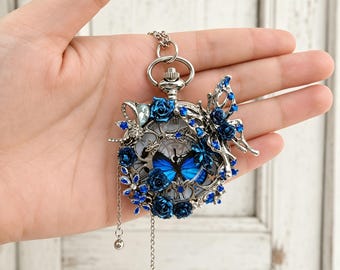 May include: A silver-toned pocket watch pendant featuring a blue butterfly motif at the center. The watch face is surrounded by intricate metal filigree, deep blue roses, and small blue crystals. A large butterfly accent sits on the right side.