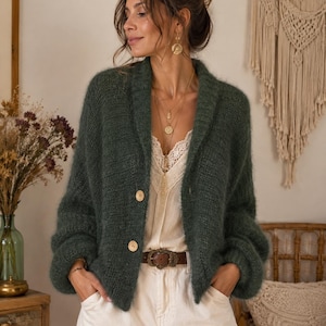 Puede incluir: Un cárdigan de punto difuso en color verde bosque oscuro con cuello de chal y dos botones grandes de madera clara. El cárdigan se lleva sobre una camisola de color crema con ribete de encaje y pantalones blancos sujetos por un cinturón de cuero.