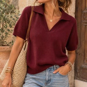 May include: A short-sleeved, deep burgundy knit polo shirt featuring a collared V-neckline and ribbed cuffs and hem. The garment is paired with light blue denim jeans and a woven straw tote bag, accessorised with a gold necklace and stacked bracelets.