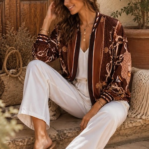 Puede incluir: Una chaqueta bomber marrón con un estampado de cadenas blancas y doradas, cierre de cremallera frontal y puños de canalé a rayas. La chaqueta se lleva sobre una camiseta blanca y se combina con pantalones blancos holgados y sandalias trenzadas.