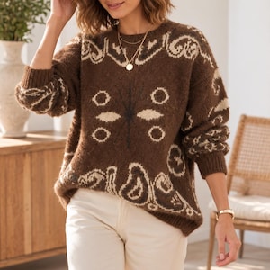 Può includere: Un maglione in maglia a maniche lunghe marrone con un motivo a farfalla color crema sul davanti e sulle maniche, caratterizzato da uno scollo rotondo e polsini a coste. Il maglione è abbinato a pantaloni color crema.
