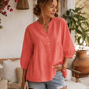 Pode incluir: Uma blusa de cor coral com decote em V, gola pequena, três botões brancos e mangas três quartos. O tecido é adornado com um delicado padrão de bordado inglês em toda a superfície e a peça tem um corte solto.