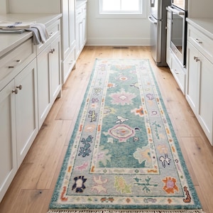 May include: A long rectangular area rug featuring a teal background with a multi-colored pattern of stylized floral and geometric motifs. The rug has a light-colored border and short fringe on both ends, positioned on a light wood floor.