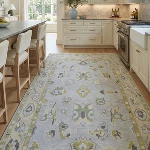Puede incluir: Una alfombra rectangular con un campo azul claro y un patrón repetitivo de motivos florales y geométricos estilizados en tonos verde salvia, crema y gris carbón, terminada con un borde decorativo y flecos cortos en los extremos.