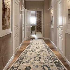 Puede incluir: Un pasillo con una larga alfombra estampada en tonos grises, verdes y crema. La alfombra recorre el suelo de madera, entre puertas y paredes blancas. Obras de arte cuelgan de las paredes y una puerta abierta revela una habitación.