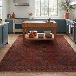 Puede incluir: Una alfombra rectangular grande con una base de color rojo intenso y patrones geométricos complejos en azul oscuro y naranja apagado. La alfombra tiene flecos decorativos en los bordes cortos y cubre el suelo en una cocina.