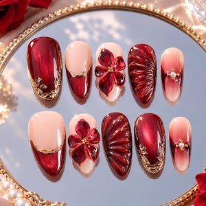 May include: A set of ten press-on nails featuring a deep red and nude color palette. The designs include solid metallic red, red floral 3D embellishments, gold filigree accents, textured fan patterns, and small white pearls set on a nude base.