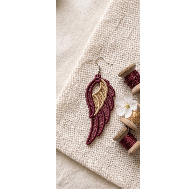 May include: An embroidered wing earring featuring deep burgundy and metallic gold thread detailing. The wing shape is constructed with dense stitching and hangs from a silver-toned metal hook. The design showcases layered feather patterns.