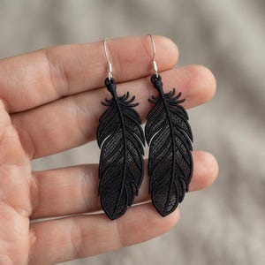 Peut inclure: Une paire de boucles d'oreilles pendantes noir mat en forme de plumes. Chaque boucle présente une structure détaillée et complexe en forme de treillis qui imite l'apparence d'une plume d'oiseau, suspendue à un crochet en métal argenté.
