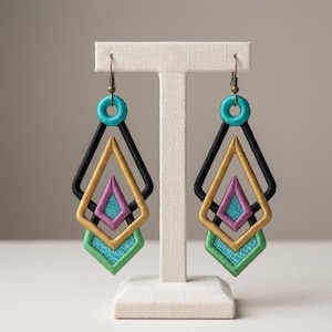 Peut inclure: Une paire de boucles d'oreilles pendantes au design géométrique superposé. Chaque boucle se compose d'un sommet circulaire turquoise, d'un cadre en losange noir, d'un cadre doré, d'un centre violet et d'une base verte, le tout brodé.