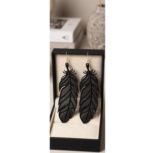 May include: A pair of black feather-shaped dangle earrings featuring a detailed lattice-like cutout pattern. The earrings are suspended from silver-toned metal hooks and are displayed inside a black rectangular jewelry box with a cream interior.