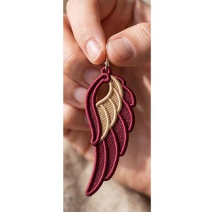 May include: A single wing-shaped earring featuring deep burgundy and metallic gold embroidery. The design consists of layered, curved segments with a prominent gold-colored inner section and a burgundy border, suspended from a silver-toned hook.