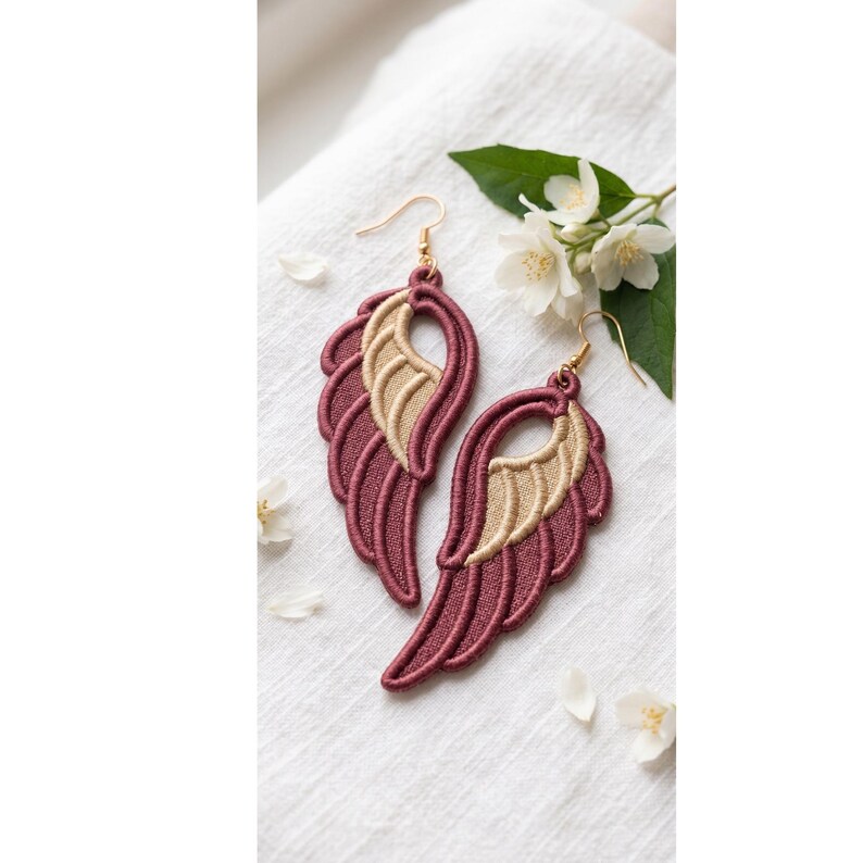 May include: A pair of embroidered wing-shaped earrings featuring a deep burgundy outer layer and a metallic gold inner section. Each earring is suspended from a gold-toned metal hook, showcasing intricate stitching details and a layered design.