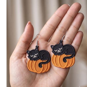 May include: A pair of dangle earrings featuring embroidered black cats sleeping on orange pumpkins. Each earring has a silver-toned metal hook for attachment. The cat and pumpkin designs are stitched with detailed thread work in black and orange.