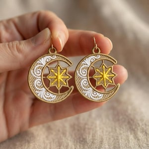 May include: A pair of circular embroidered earrings featuring a crescent moon shape in tan thread with white swirling patterns. Centered within each moon is a bright yellow eight-pointed star. The earrings hang from small gold-toned metal hooks.