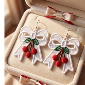 May include: A pair of dangle earrings featuring white embroidered bows with green leaves and three small red cherries hanging from the center. The earrings have silver-toned metal hooks and are displayed inside a cream-colored jewelry box.