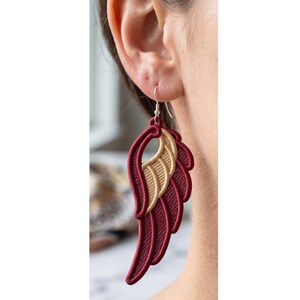 May include: A dangling earring shaped like a stylized wing, featuring deep burgundy embroidery for the outer feathers and a contrasting metallic gold thread for the inner wing section, suspended from a simple silver-toned metal hook wire.