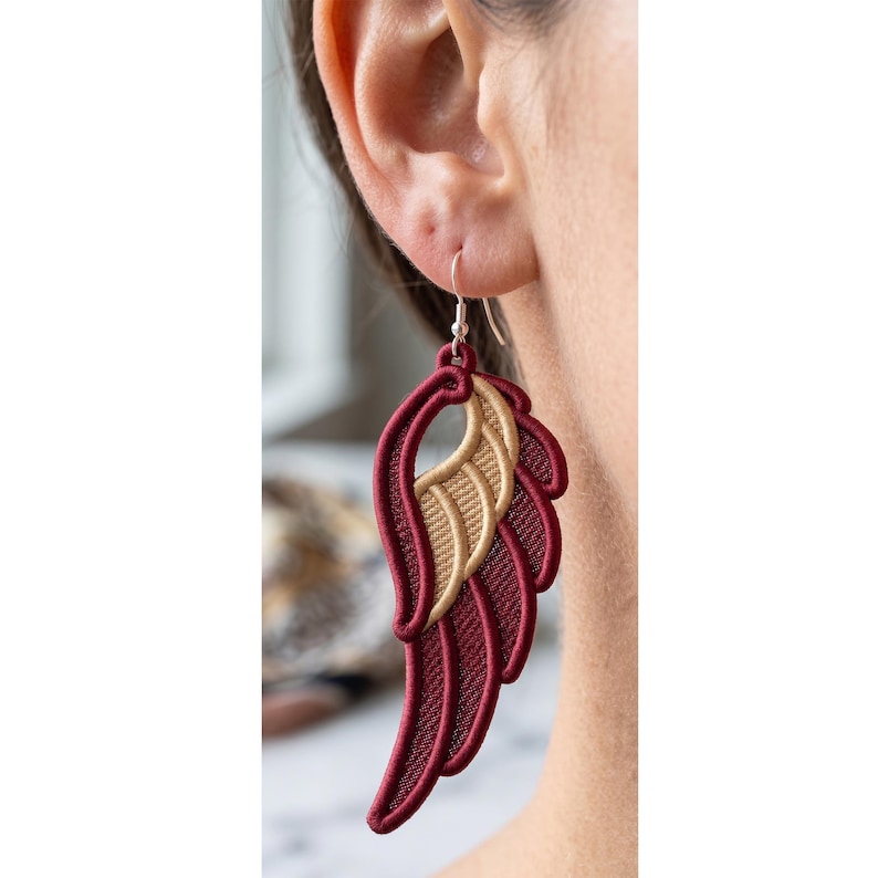 May include: A dangling earring shaped like a stylized wing, featuring deep burgundy embroidery for the outer feathers and a contrasting metallic gold thread for the inner wing section, suspended from a simple silver-toned metal hook wire.