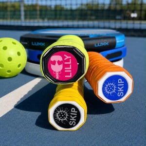 May include: Three pickleball paddles with custom end caps and a neon yellow pickleball. One cap features a pink design with a heart-eyed paddle icon and the name LILLY. Two caps feature a blue or black graphic of a pickleball and the name SKIP.