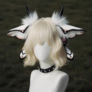 Op de afbeelding: Een witte pruik op een etalagepophoofd met grote motachtige oren van wit imitatiebont, zwarte randen en kleine zilveren sterren. Twee zwarte hoorns steken uit en een zwarte halsband met studs en een centrale metalen ring zit om de nek.