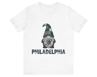 May include: A white short-sleeve t-shirt featuring a graphic of a gnome wearing a plaid hat and holding a football. The gnome is positioned above the text PHILADELPHIA, which is written in a dark green font with a plaid pattern fill.