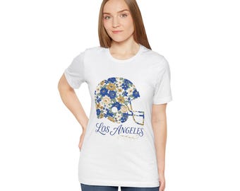 May include: A white short-sleeve t-shirt featuring a graphic of a football helmet filled with a blue, gold, and white floral pattern. Below the helmet, the text Los Angeles is printed in a blue script font with a small decorative flourish.