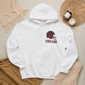 Puede incluir: Una sudadera con capucha blanca que presenta un gráfico en el pecho de un casco de fútbol americano con un patrón de cuadros azul marino y naranja. Debajo del casco, la palabra CHICAGO está impresa en letras azul marino y naranja.