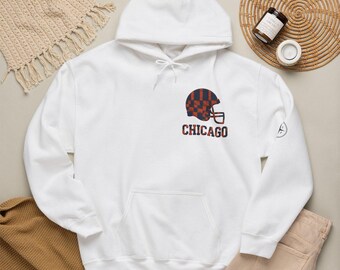 May include: A white hooded sweatshirt featuring a graphic on the chest of a football helmet with a navy and orange checkered pattern. Below the helmet, the word CHICAGO is printed in navy and orange letters. The hoodie has a front kangaroo pocket.