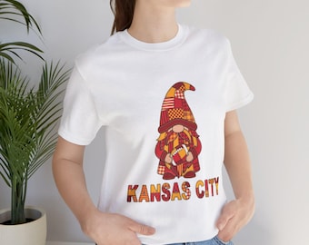 May include: A white short-sleeve t-shirt featuring a graphic of a gnome wearing a red and orange plaid hat and outfit, holding a football. Below the gnome, the text KANSAS CITY is printed in a matching red and orange plaid pattern.