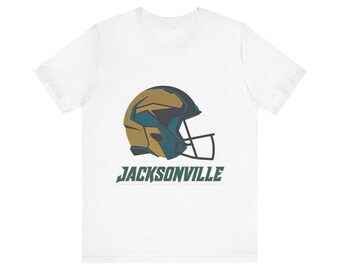 Könnte beinhalten: Ein weißes Kurzarm-T-Shirt mit einer Grafik eines Football-Helms in Gold-, Petrol- und Dunkelgrautönen. Unter dem Helm ist das Wort JACKSONVILLE in einer fetten, dunkelpetrolfarbenen, serifenlosen Schrift mit einer leichten kursiven Neigung gedruckt.