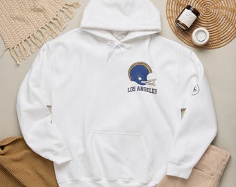 May include: A white hooded sweatshirt featuring a graphic on the chest of a blue and gold football helmet above the text LOS ANGELES. The hoodie has long sleeves, a front kangaroo pocket, and a drawstring at the neck with white cords.