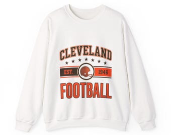 Könnte beinhalten: Ein weißes langärmeliges Sweatshirt mit Rundhalsausschnitt und einem Grafikdruck, der CLEVELAND FOOTBALL in orangefarbenen und schwarzen Buchstaben zeigt. Das Design enthält ein braunes Football-Helm-Symbol, den Text EST. 1946 und sieben schwarze Sterne.