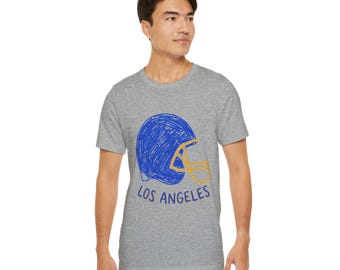 May include: A heather grey short-sleeve t-shirt featuring a blue and yellow scribbled graphic of a football helmet. Below the helmet, the text LOS ANGELES is printed in blue. The design has a hand-drawn, sketch-like appearance on the fabric.