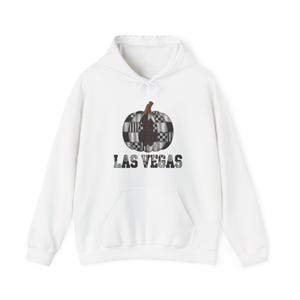 Op de afbeelding: Een witte hoodie met een centrale afbeelding van een pompoen met een zwart-wit geblokt patroon. Onder de pompoen staat de tekst LAS VEGAS gedrukt in een vet, verweerd zwart lettertype. De hoodie is voorzien van een kangoeroezak aan de voorzijde.