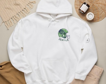 Könnte beinhalten: Ein weißer Kapuzenpullover mit einer Kängurutasche und weißen Kordelzügen. Auf der Brust befindet sich eine skizzenartige Grafik eines Football-Helms in Grün und Blau, unter der das Wort SEATTLE in dunkelgrünen Buchstaben gedruckt ist.