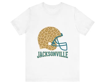 May include: A white short-sleeve t-shirt featuring a graphic of a football helmet with a tan and brown leopard print pattern and a teal face mask. Below the helmet, the word JACKSONVILLE is printed in a teal, textured, block-style font.