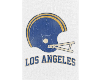 Puede incluir: Una toalla rectangular blanca con un gráfico de un casco de fútbol americano azul y amarillo desgastado con una máscara blanca. Debajo del casco, el texto LOS ANGELES está impreso en una fuente azul, arqueada y con borde amarillo.